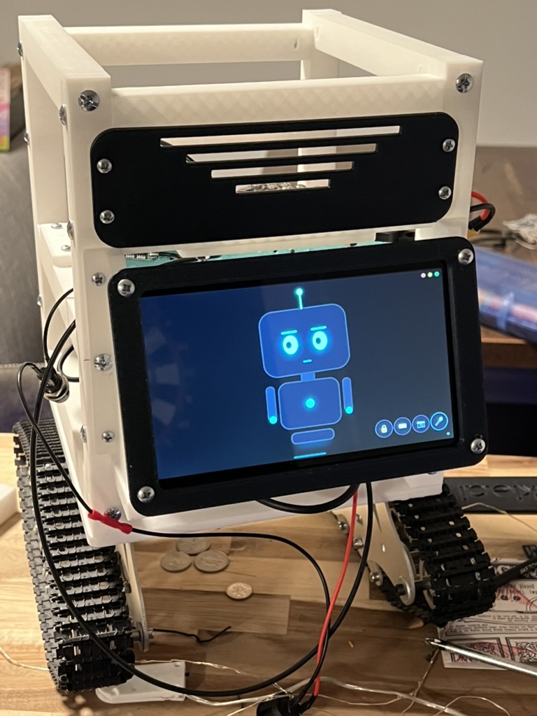 The v3 Meckie prototype on the workbench: white printed frame, charcoal grille panel, belly screen on and showing Meckie's blue robot face, tracks underneath, loose wires and a soldering iron on the bench.