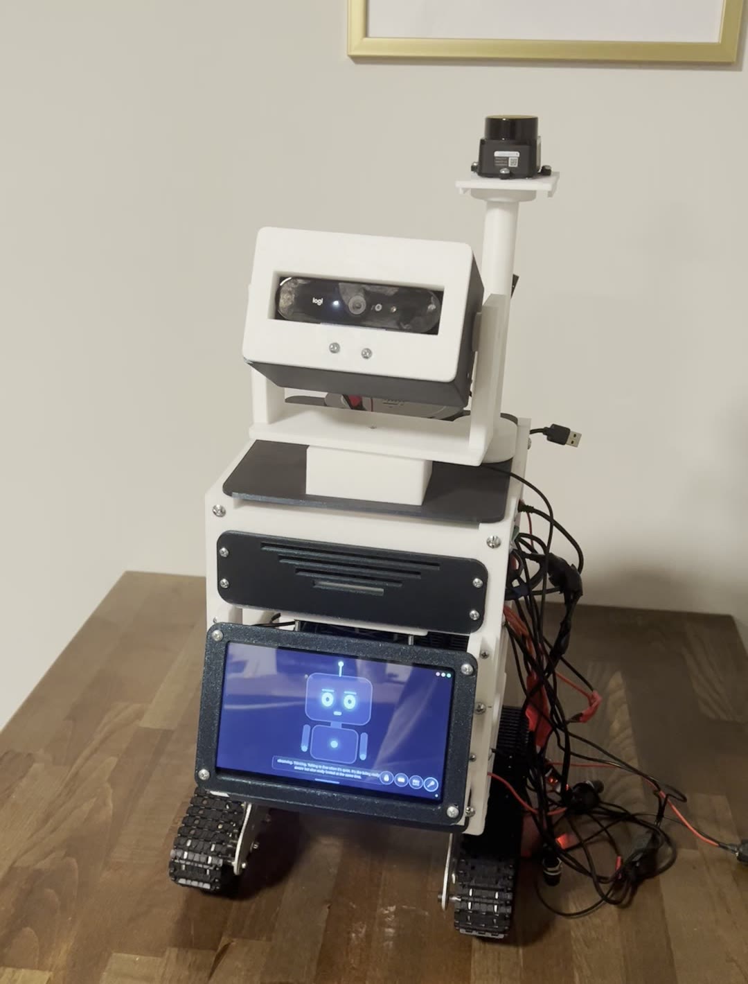 Droid on the workbench with his head turned to the side: white printed body on tank tracks, LiDAR up top, camera head swiveled mid-glance, belly screen showing his blue face.