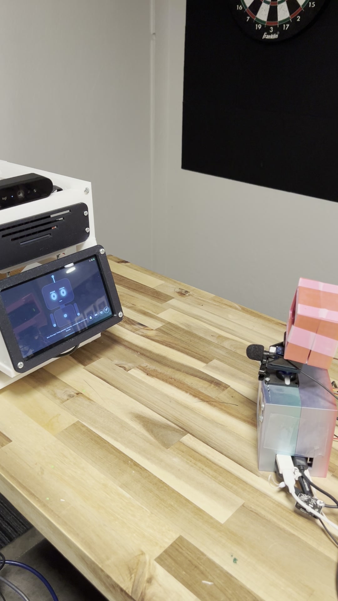 Two Meckie robots facing each other on a wood workbench: Droid, the v3 build with his face on the belly screen, and Eva, a smaller Pi robot with a pink top and a little microphone, dartboard on the wall behind them.