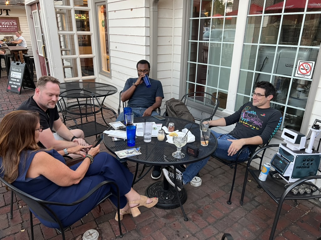 Four friends around a patio table at Ziggy's Pizza in Wichita at golden hour, with Droid the robot parked at the table's edge and a small speaker on the table