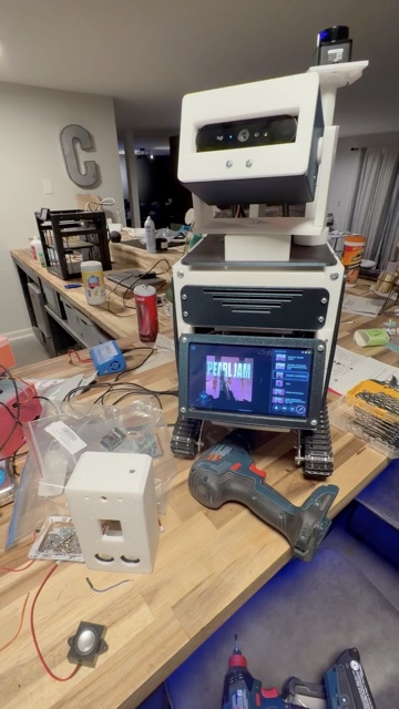 A workbench mid-build: a small white 3D-printed robot shell in the foreground next to a drill and a bare speaker driver, with Droid the tracked robot standing behind it playing Pearl Jam on his belly screen