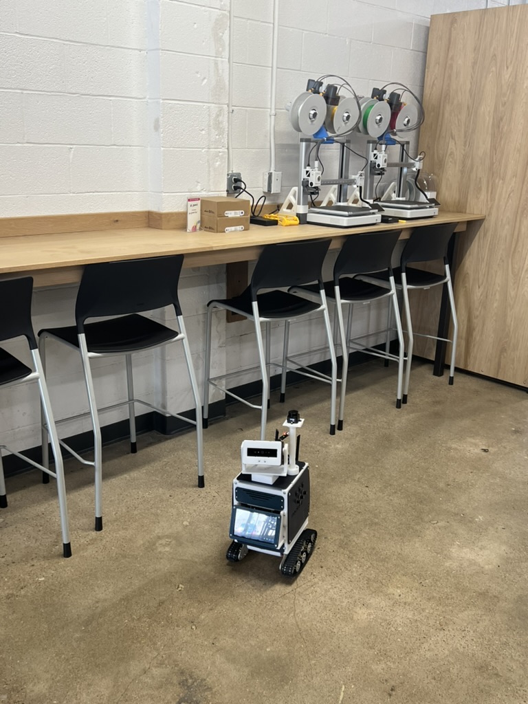 Droid, a small tracked robot with a lidar mast and belly screen, parked on the polished concrete floor of Groover Labs in front of a workbench lined with 3D printers