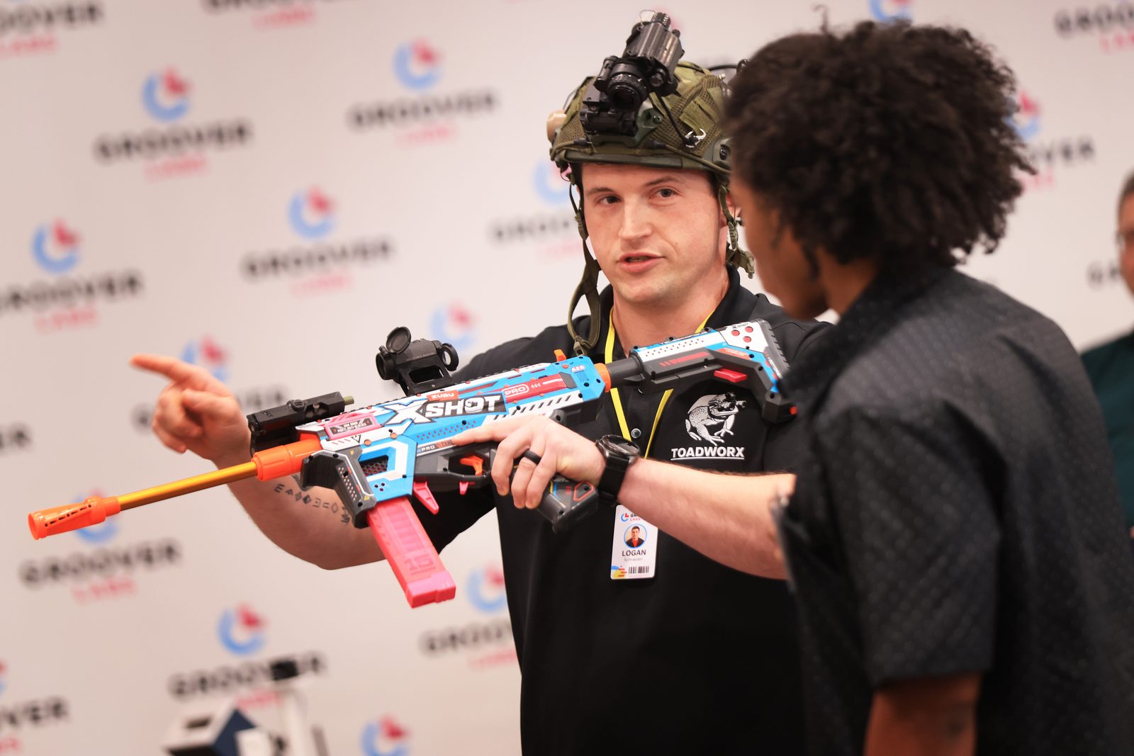 A ToadWorx presenter in a tactical helmet and night-vision mount handing a nerf gun to an eager young man.