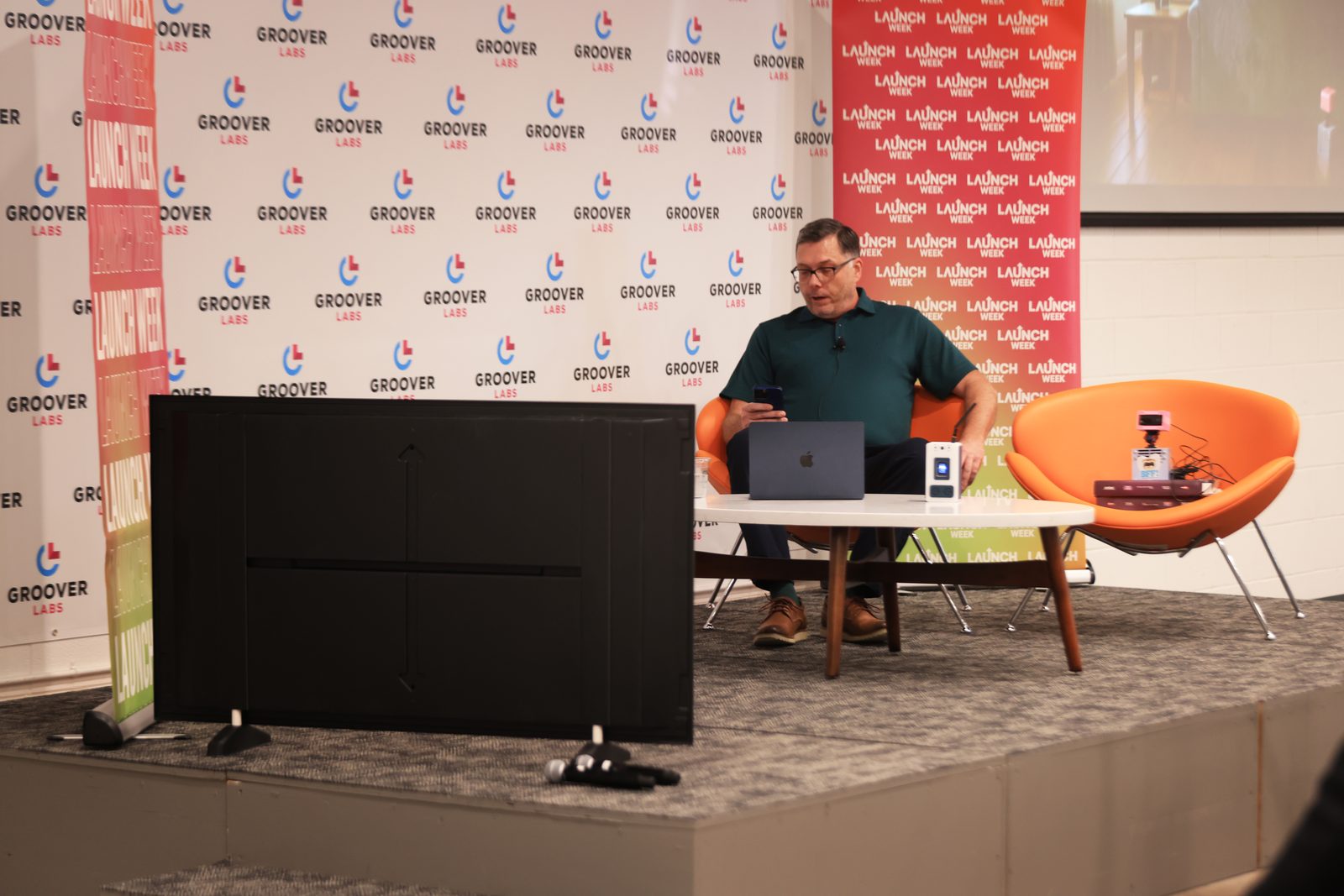 Chad on stage at Groover Labs during Launch Week, a laptop and a Meckie on the table, a large display propped on its side in front of the stage.