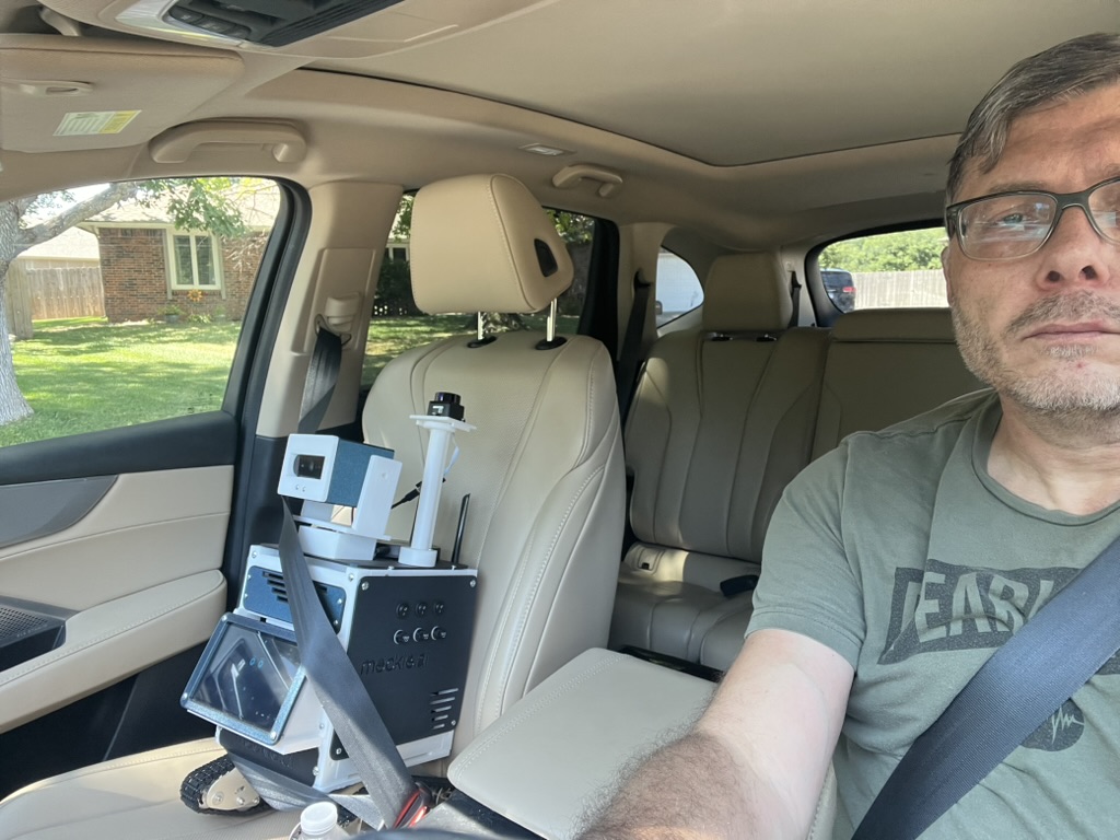 Droid the Meckie buckled into the passenger seat of a car with the seatbelt across his chassis, camera head upright, on his way to map a venue